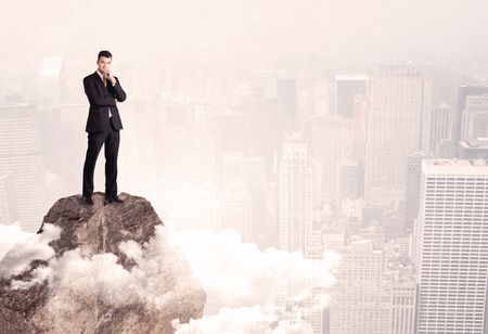 An elegant professional business male standing on top of a high cliff above the clouds looking at the city concept