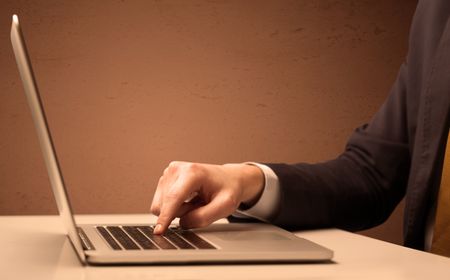 An office worker in elegant suit sitting at desk, typing on portable laptop with empty brown wall background