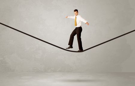 An elegan businessman in suit balancing on a tight rope with a briefcase in front of grey urban wall background environment concept