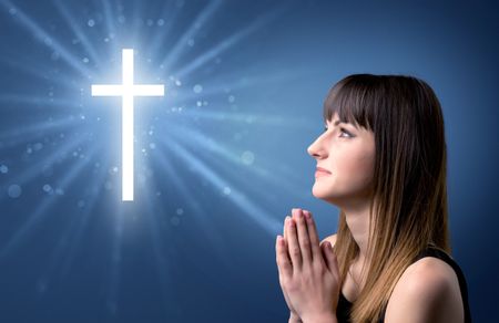 Young woman praying on a blue background with a sparkling cross above her