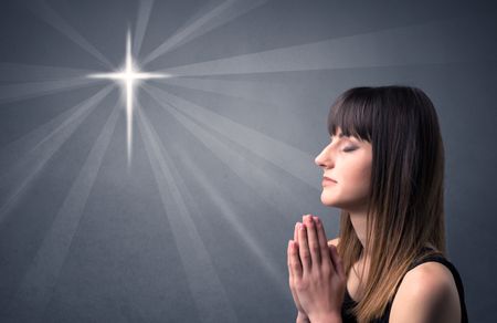 Young woman praying on a grey background with a shiny cross silhouette above her