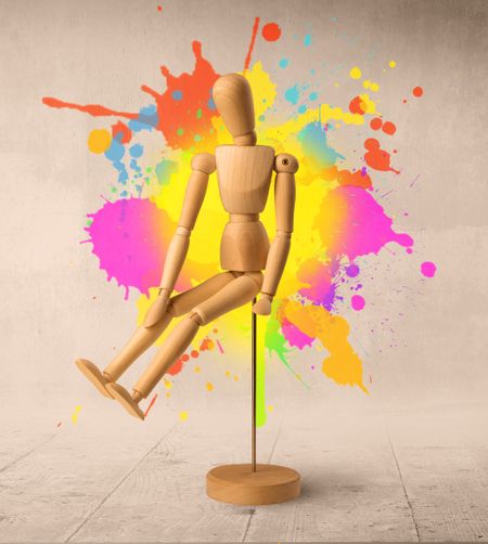 Wooden mannequin posed in front of a greyish background with colorful splashes behind it