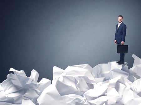 Thoughtful young businessman standing on a pile of crumpled paper with a blueish grey background