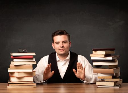 A young teacher in glasses sitting at classroom desk with pile of books in front of clean blackboard back to school concept.