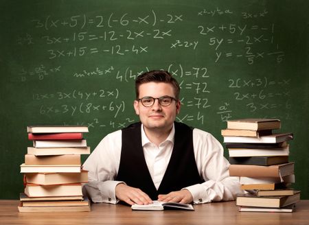 A young ambitious teacher in glasses sitting at classroom desk with pile of books in front of blackboard full of math calculations, numbers, back to school concept.