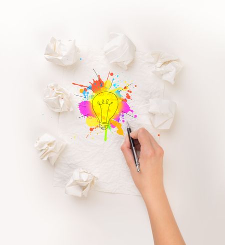 Female hand next to a few crumpled paper balls drawing a colorful lightbulb