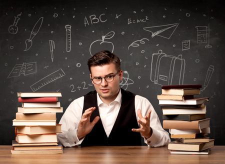 A passionate young teacher sitting at school desk with pile of books in front of blackboard drawn full of back to school items concept.