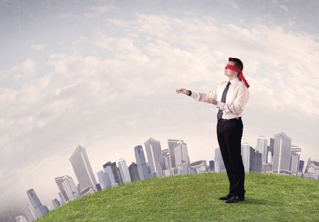 Young blindfolded businessman steps on a a patch of grass with a grey buildings in the background