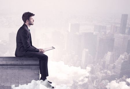 An elegant young businessman sitting on the edge looking over urban city landscape with clouds concept