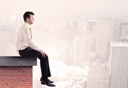 An elegant businessman in modern suit sitting on the top of a brick building, looking over the cityscape with clouds concept