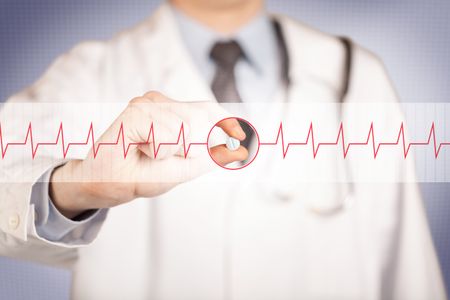 A male doctor in white coat with a stethoscope on one shoulder holding a pill between his fingers focused on a heartbeat graph