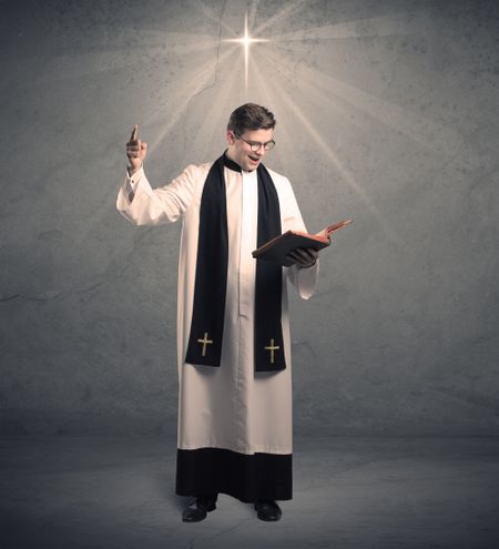 A young male priest in black and white giving his blessing in front of grey wall with glowing cross concept.