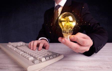 An elegant office worker holding a yellow sparkling light bulb in his hand while working in front of dark blue background concept.