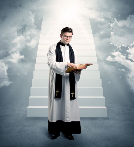 A young religious happy priest standing in front of the stairway to heaven concept with clouds and bright lights coming from above.