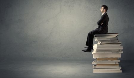 A serious student in elegant suit sitting on a stack of books in front of dark grey background