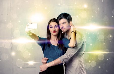 A fresh happy couple taking selfie photo with mobile phone in front of colorful lights glitter wall background concept