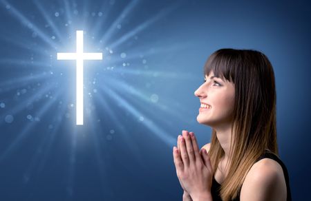 Young woman praying on a blue background with a sparkling cross above her