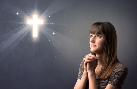 Young woman praying on a grey background with a shiny cross above her