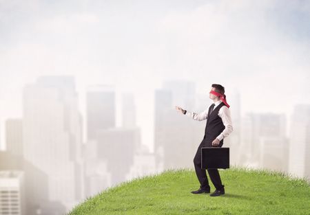 Young blindfolded businessman steps on a a patch of grass with a city in the background 