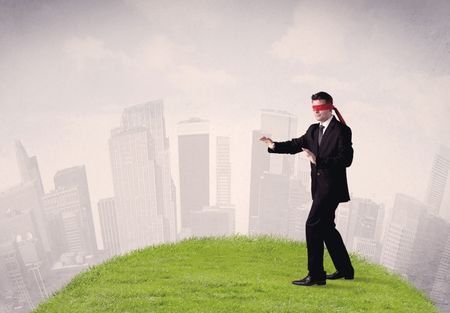 Young blindfolded businessman steps on a a patch of grass with a cloudy city in the background