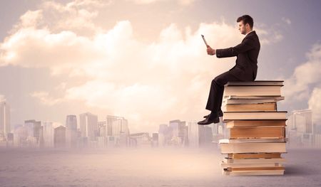 A serious student with laptop tablet in elegant suit sitting on a stack of books in front of cityscape with clouds