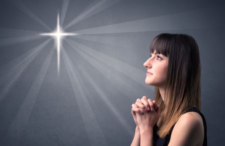 Young woman praying on a grey background with a shiny cross silhouette above her