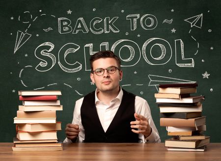 A young teacher in glasses sitting at classroom desk with pile of books in front of blackboard saying back to school drawing concept.