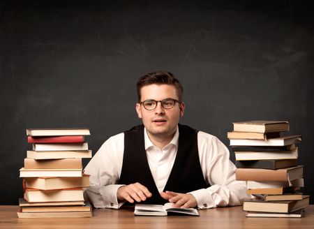 A young teacher in glasses sitting at classroom desk with pile of books in front of clean blackboard back to school concept.