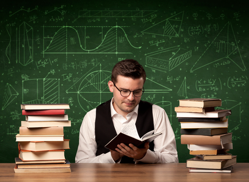 A young passionate male teacher sitting at school desk, reading a book, with area algorythm calculations and numbers on the blackboard concept.