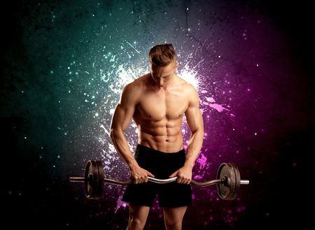 A sexy male fitness trainer showing his muscles and looking seductive with a weight in his hands in front of bright paint splash purple wall concept