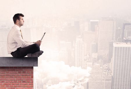 An elegant businessman in modern suit sitting on the top of a brick building, looking over the cityscape with clouds concept