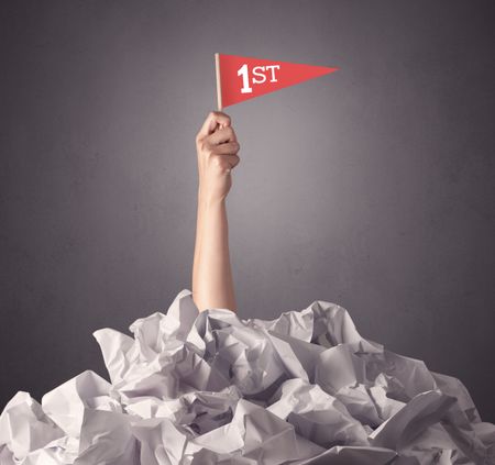 Female hand emerging from crumpled paper pile holding a red flag with first written on it