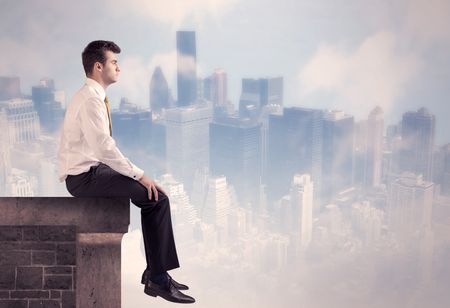 A young businessman sitting at the edge of a building in front of a city center scape background with tall buildings and clouds concept