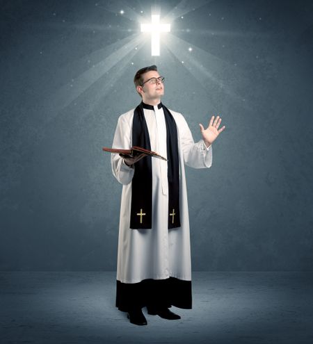 A young caucasian priest with deep faith blessing with the holy bible in his hand in front of blue wall background with illustrated glowing cross concept.