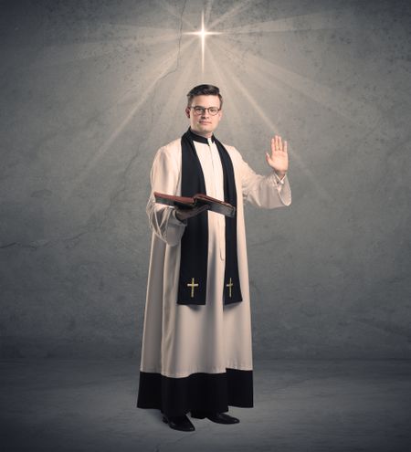A young male priest in black and white giving his blessing in front of grey wall with glowing cross concept.