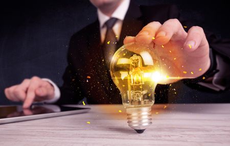 An elegant office worker holding a yellow sparkling light bulb in his hand while working in front of dark blue background concept.