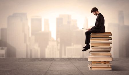 A serious businessman with laptop tablet in elegant suit sitting on a stack of books in front of cityscape
