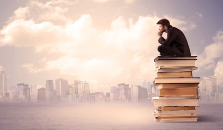 A serious businessman in elegant suit sitting on a stack of books in front of city scape and clouds