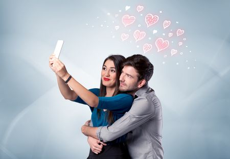 A young couple in love and drawn red hearts taking selfie with a mobile phone in the handsome guy's hand in front of an empty clear grey wall background concept