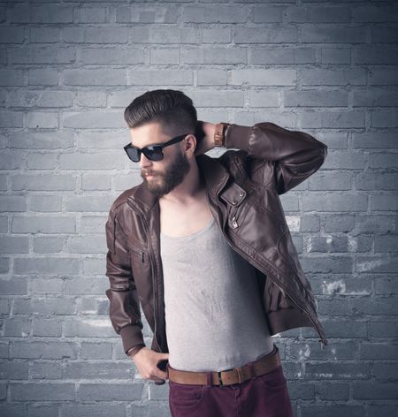 A stylish hipster guy with beard and sunglasses standing in casual clothes in front of an urban blue brick wall background concept