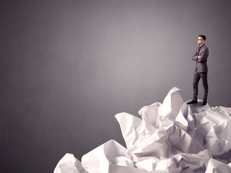 Thoughtful young businessman standing on a pile of crumpled paper with a grungy grey background