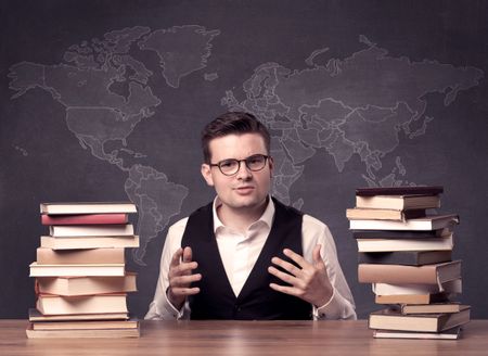 A young ambitious geography teacher in glasses sitting at classroom desk with pile of books in front of world map drawing on blackboard, back to school concept.