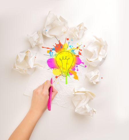 Female hand next to a few crumpled paper balls drawing a colorful lightbulb