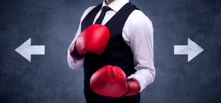 A confident businessman standing with red boxing gloves on his hand in front of arrows pointing in different directions on urban wall background concept.