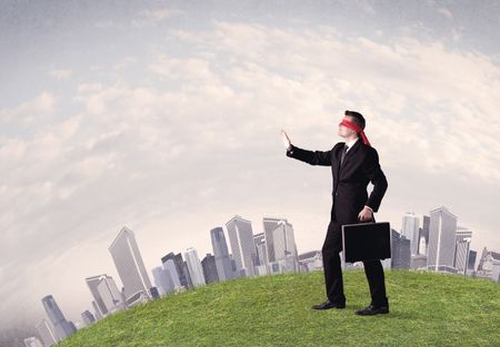 Young blindfolded businessman steps on a a patch of grass with a grey buildings in the background