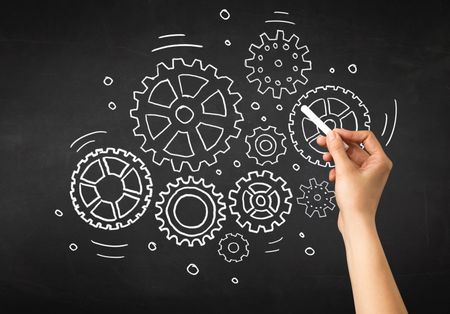 Female hand holding white chalk in front of a blackboard with gears drawn on it