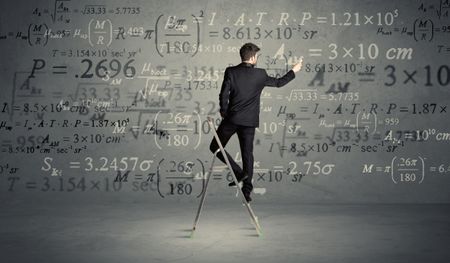 A businessman in elegant suit standing on a small ladder and writing numbers, calculating on grey wall background