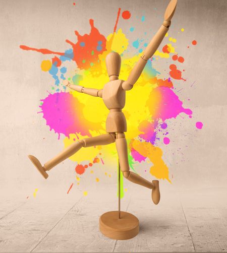 Wooden mannequin posed in front of a greyish background with colorful splashes behind it