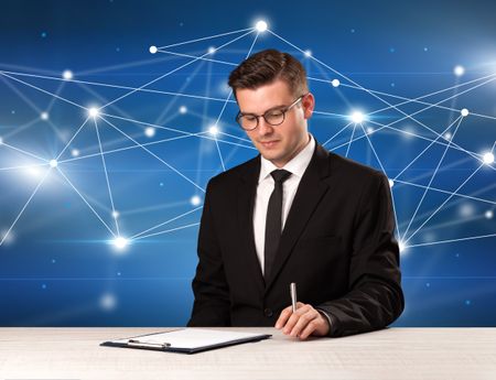 Young handsome businessman sitting at a desk with a blue connection graphic behnid him 