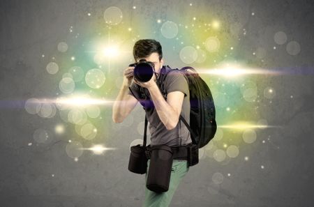 A young amateur photographer with professional camera equipment taking picture in front of grey wall full of colorful bokeh and glowing lights concept